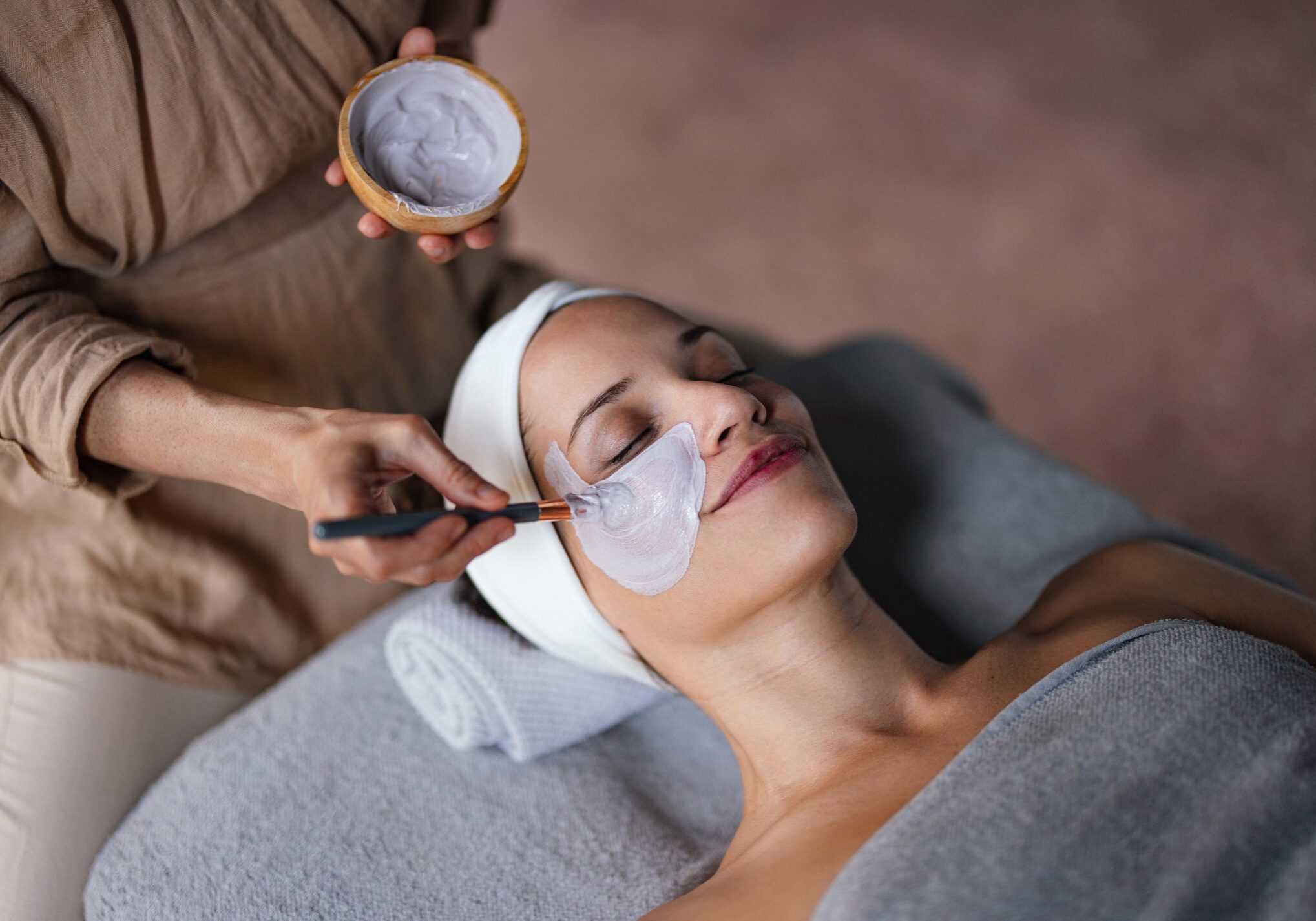 Close up shot of a beautiful smiling woman lying with her eyes closed while another woman is applying a face mask on her in the spa center. She is relaxed and enjoys the treatment.